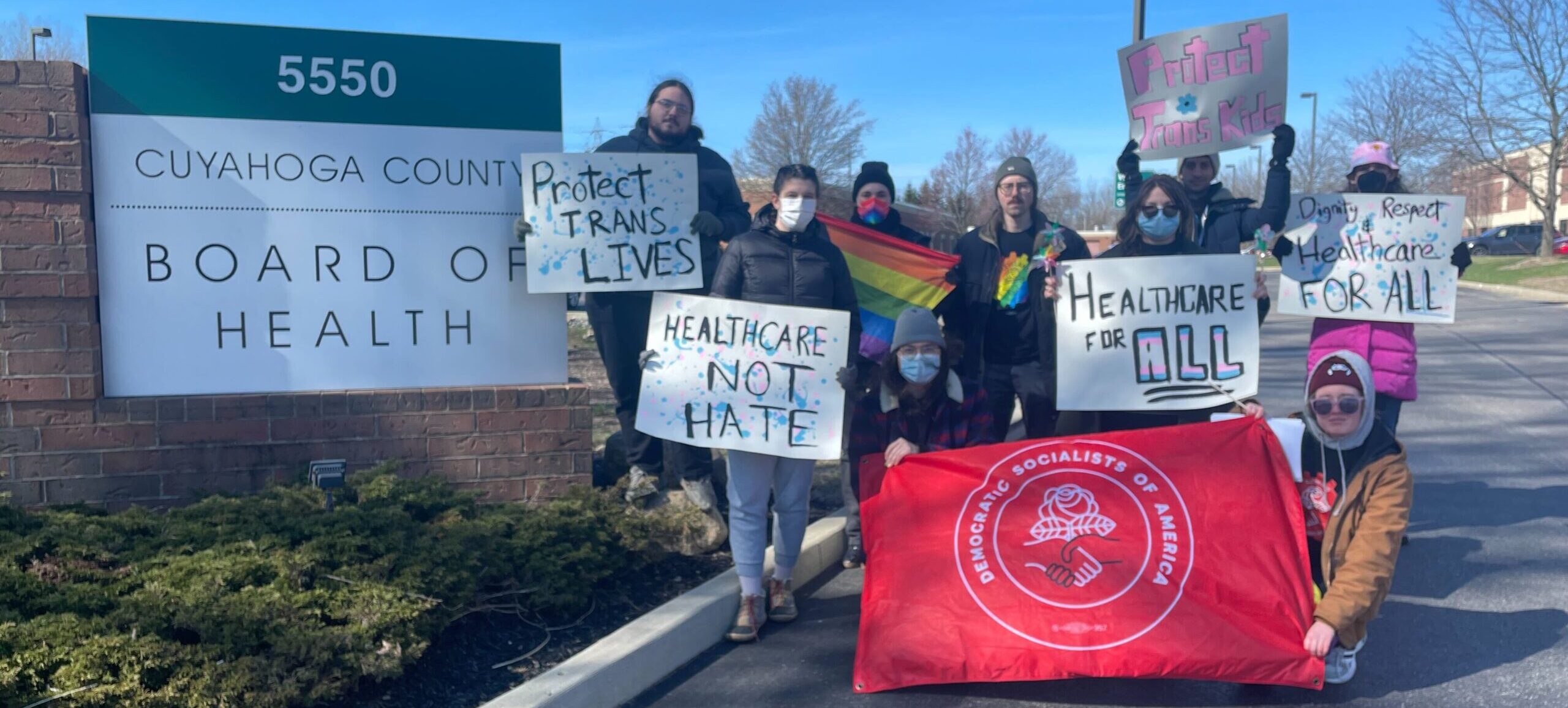 Trans Activists Post with Signs and a DSA Cleveland Flag at the Ohio Department of Health Meeting Where Public Comment Was Made Against the Anti-Trans Administrative Rules