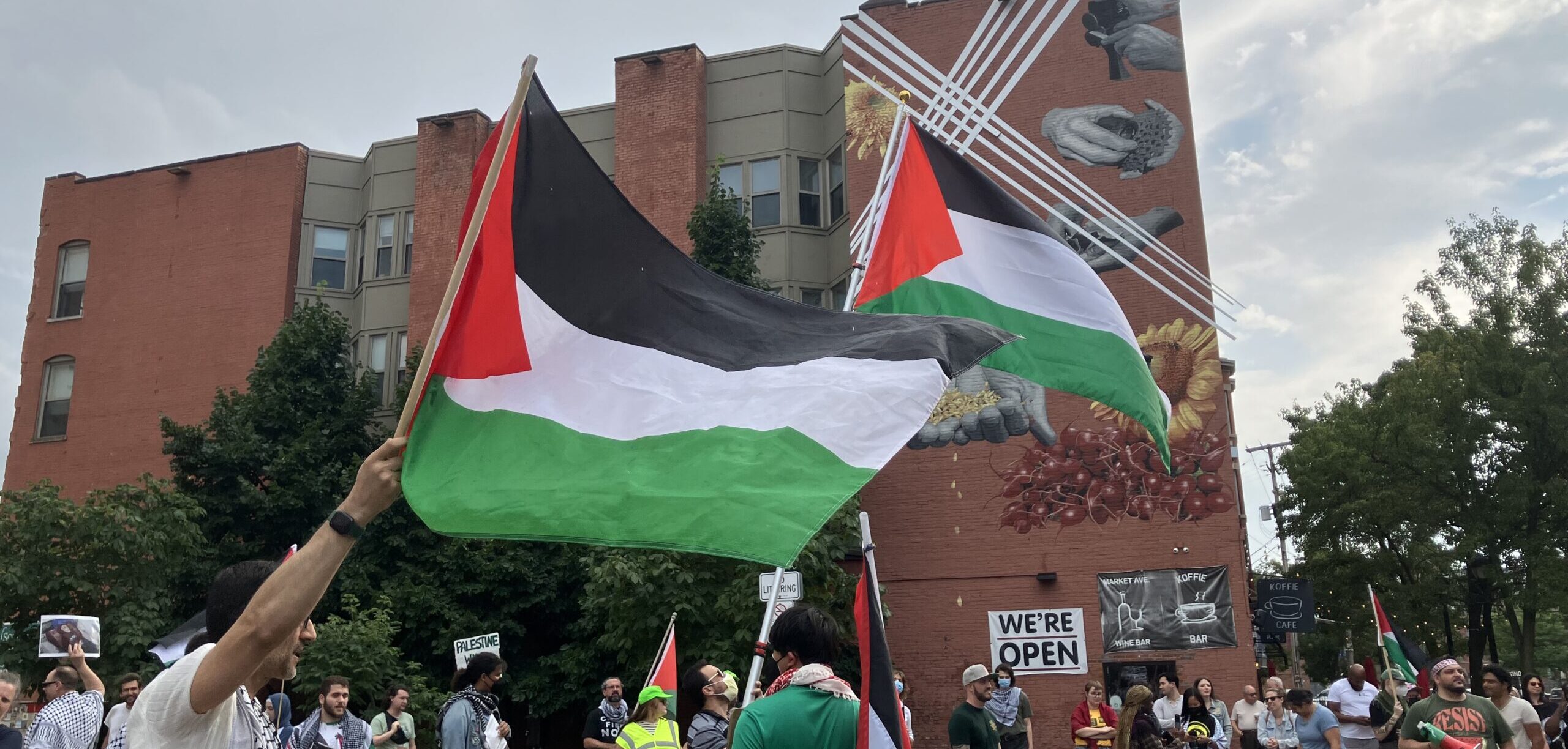 Photo of a pro-Palestine rally at Market Square in Downtown Cleveland. Two large Palestinian flags are being waved high in the foreground, with supporters in the background.